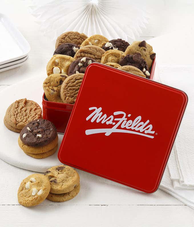  Red Mrs. Fields tin filled with a variety of mini cookies, including chocolate chip, white chocolate macadamia, and double chocolate, displayed with glasses of milk and striped straws on a white wooden surface with a red checkered napkin.
