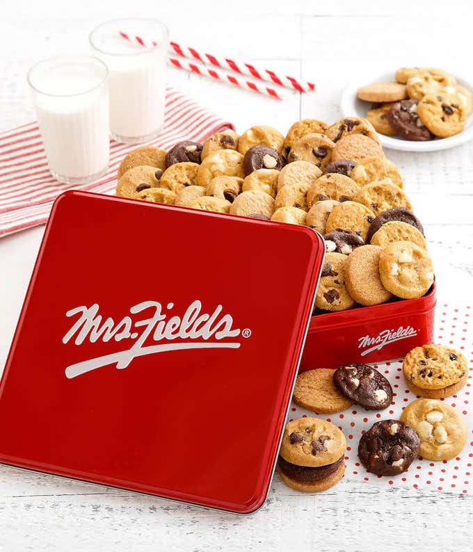  Red Mrs. Fields tin filled with an assortment of mini cookies, including chocolate chip, white chocolate macadamia, and double chocolate, arranged on a white wooden surface with glasses of milk and striped straws in the background.