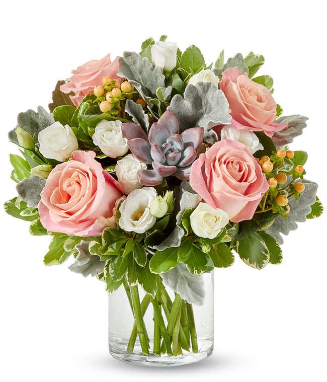 Floral arrangement in a clear glass vase featuring soft pink roses, white spray roses, peach hypericum berries, silver dusty miller, green foliage, and a central mauve-toned succulent.