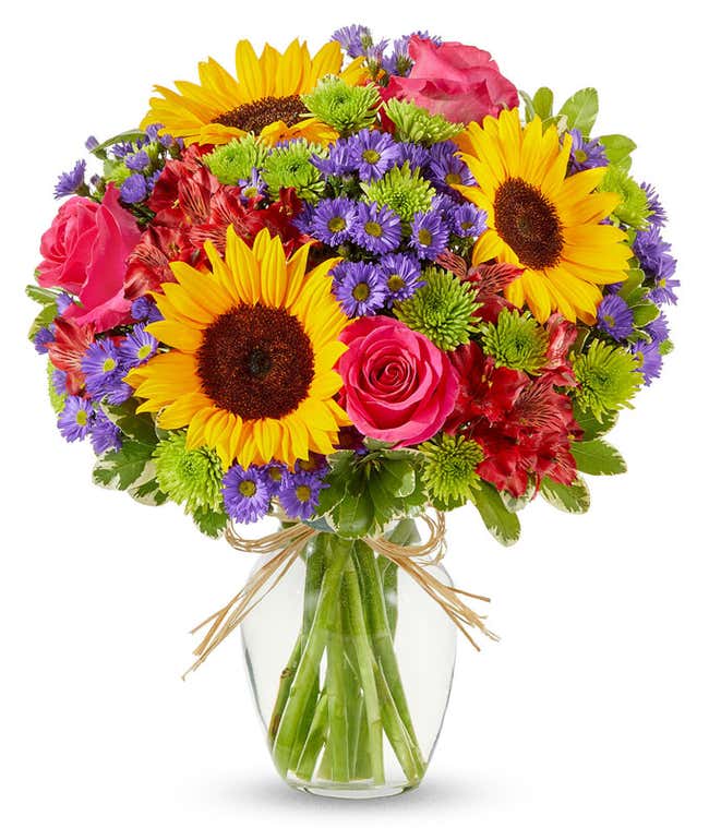 Bright mixed flower bouquet featuring yellow sunflowers, hot pink roses, red alstroemeria, green button poms, and purple asters in a clear glass vase tied with raffia ribbon.