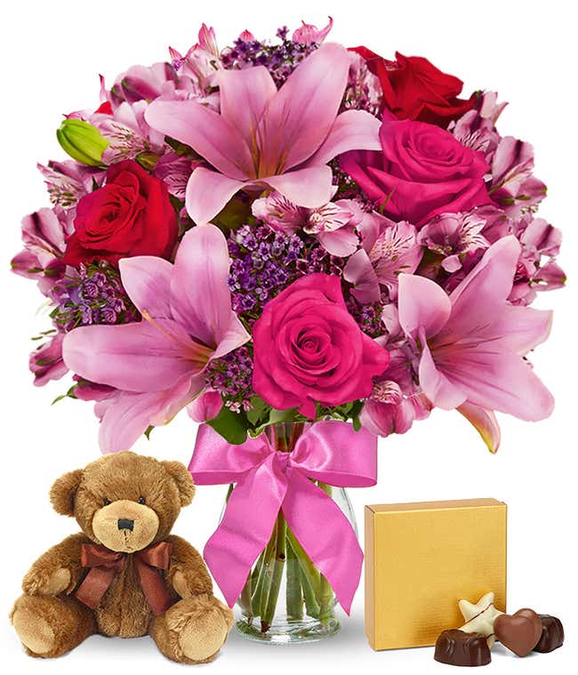 Romantic gift set with a bouquet of pink lilies, hot pink and red roses, pink alstroemeria, and purple filler in a glass vase with a pink bow, paired with a plush teddy bear and box of assorted chocolates. 