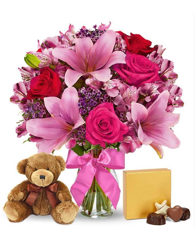 Romantic gift set with a bouquet of pink lilies, hot pink and red roses, pink alstroemeria, and purple filler in a glass vase with a pink bow, paired with a plush teddy bear and box of assorted chocolates. 