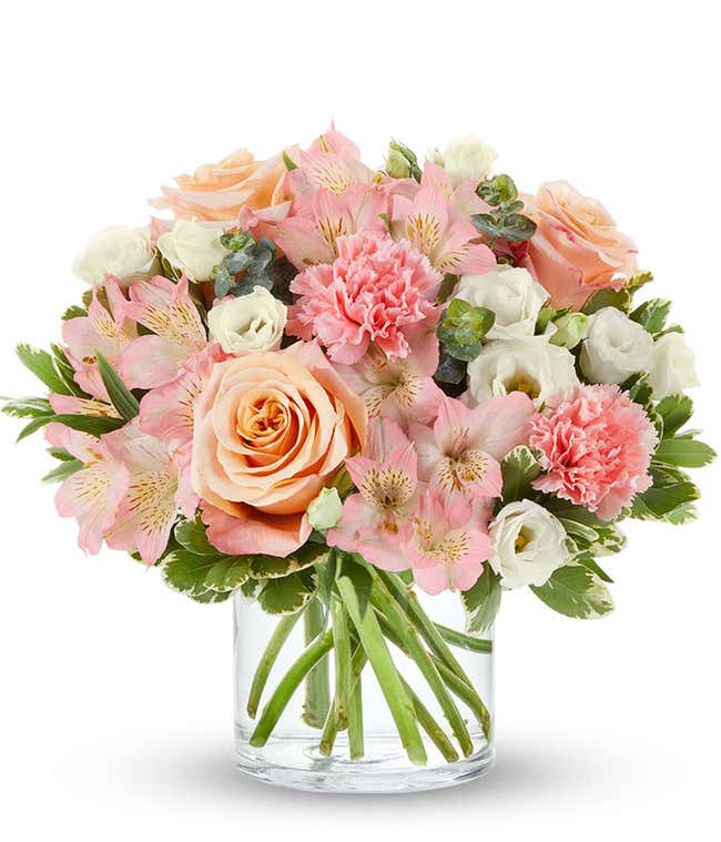 Bouquet of peach roses, pink carnations, pink alstroemeria, and white lisianthus with greenery, arranged in a clear glass cylinder vase.