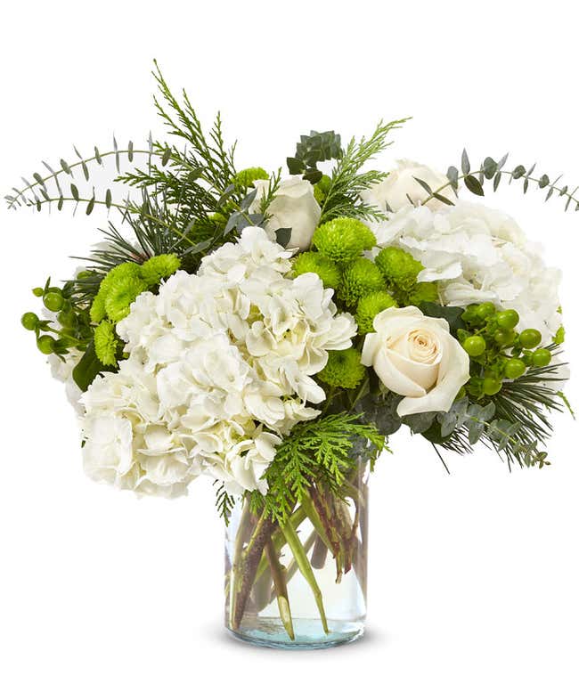 Bouquet of white hydrangeas, white roses, green button poms, green hypericum berries, and assorted evergreen foliage arranged in a clear glass cylinder vase.