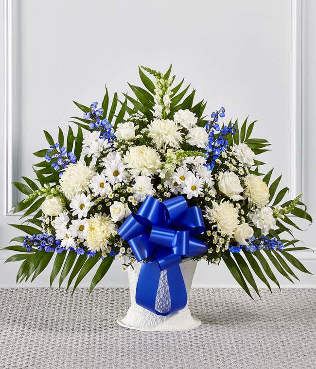 Standing floral arrangement of white roses, chrysanthemums, carnations, daisies, and snapdragons accented with blue delphinium and a large royal blue ribbon bow.