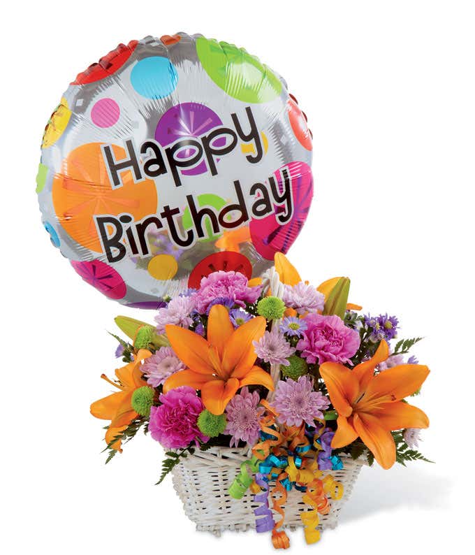 Festive birthday flower arrangement with orange lilies, pink carnations, purple poms, green button poms, and a colorful “Happy Birthday” balloon in a white basket.