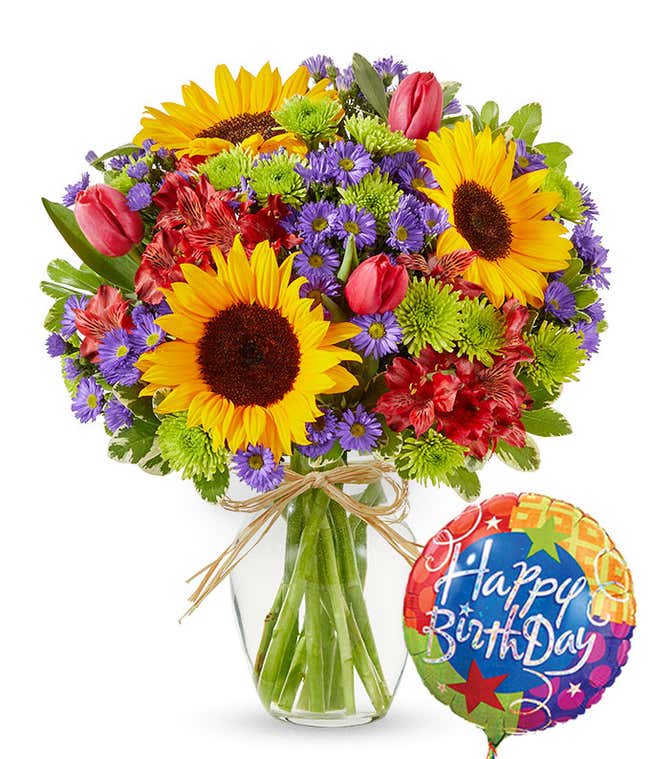 Clear glass vase filled with sunflowers, red alstroemeria, pink tulips, green button poms, and purple asters, tied with raffia and paired with a colorful Happy Birthday balloon.