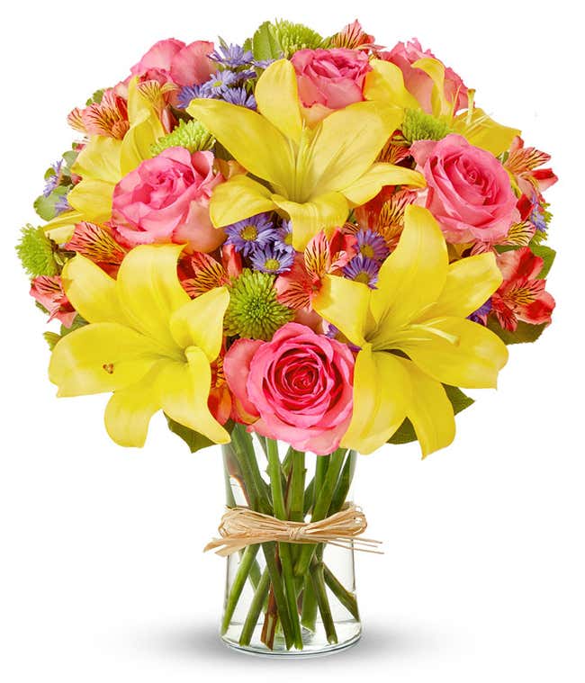 A vibrant arrangement featuring yellow lilies, pink roses, pink alstroemeria, green poms, and purple Monte Casino, beautifully arranged in a glass vase.