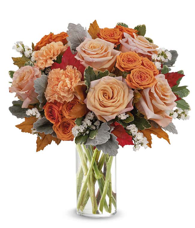 Clear glass vase filled with a lush arrangement of peach and orange roses, orange carnations, white filler flowers, dusty miller, red and orange autumn leaves, and assorted greenery.