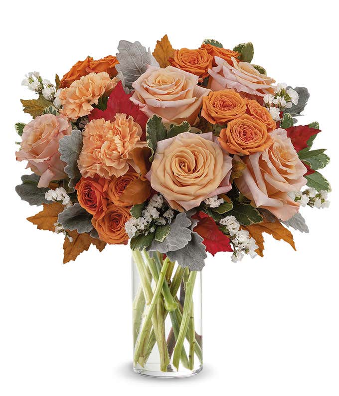 Clear glass vase filled with a lush arrangement of peach and orange roses, orange carnations, white filler flowers, dusty miller, red and orange autumn leaves, and assorted greenery.