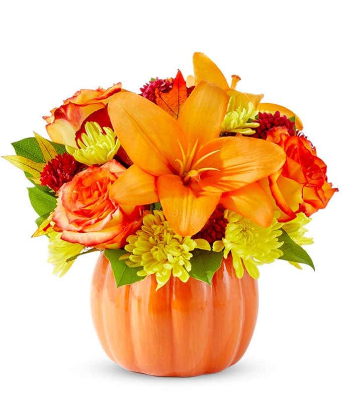 Bright fall flower arrangement with orange lilies, orange roses, yellow poms, red accent blooms, and greenery in a ceramic pumpkin container.