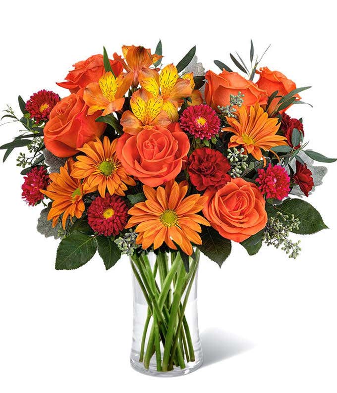 A vibrant floral arrangement featuring orange roses, orange alstroemeria, bronze daisies, red Matsumoto asters, maroon mini carnations, and seasonal greenery, complemented by a personal card message, all beautifully presented in a clear cylinder vase.