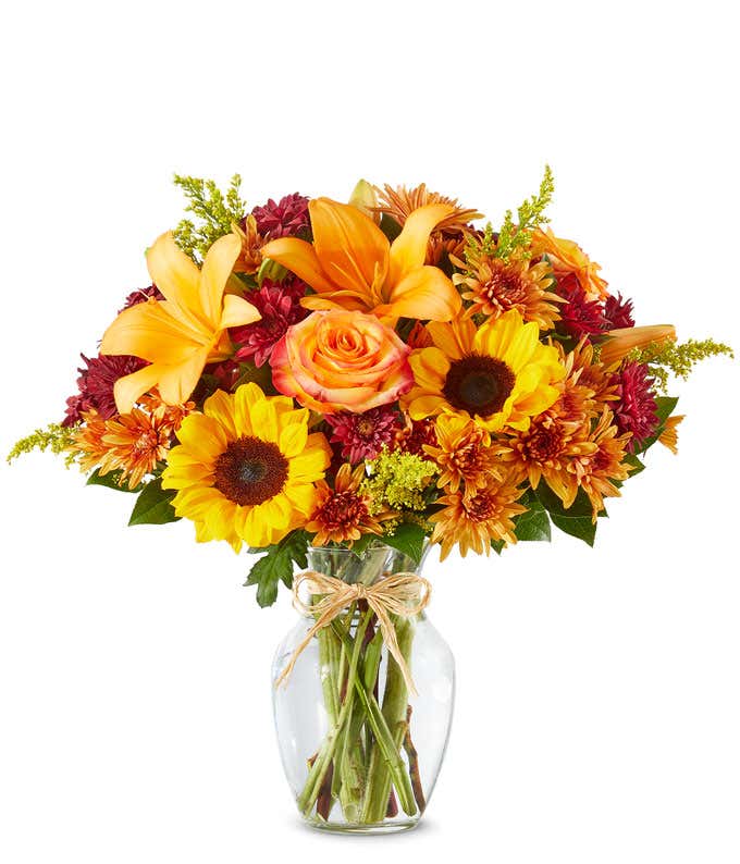 Bright autumn bouquet with sunflowers, orange lilies, orange roses, burgundy poms, yellow solidago, and butterscotch daisies in a clear glass vase tied with a raffia bow.