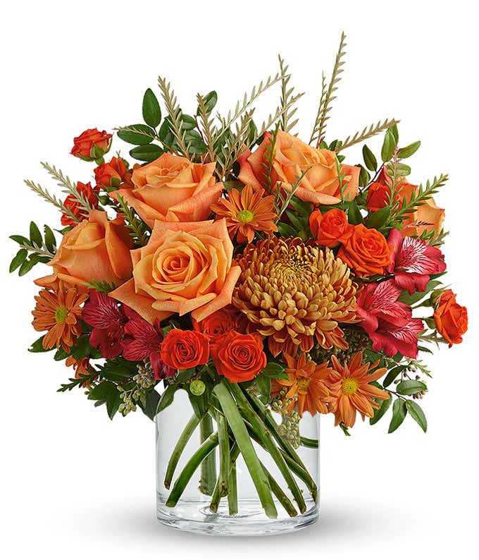 Autumn floral arrangement with orange roses, bronze daisy poms, red alstroemeria, golden mum, orange spray roses, and greenery in a clear glass cylinder vase.