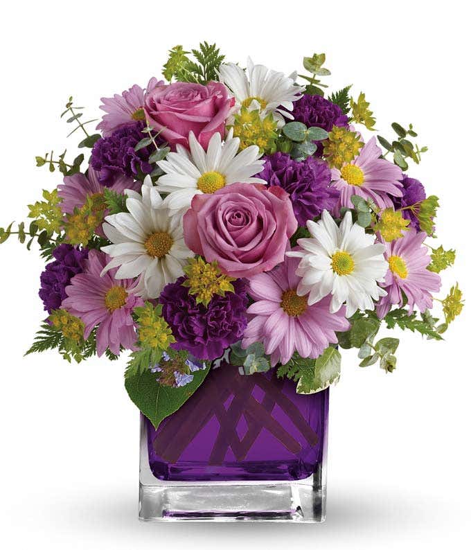 A beautiful arrangement featuring purple carnations, lavender daisy spray mums, white daisy spray mums, bupleurum, spiral eucalyptus, and leatherleaf fern, presented in a purple glass cube vase.