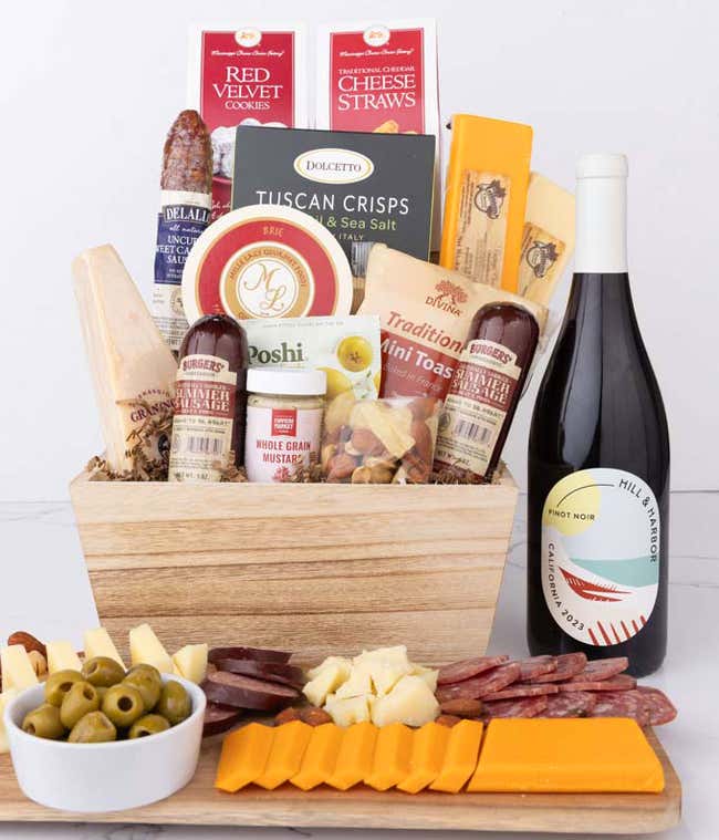 Wooden basket filled with 4 cheeses, 3 sausages, crackers, cookies, nuts, mustard, olives and crisps alongside a bottle of Hill &amp; Harbor Pinot Noir.