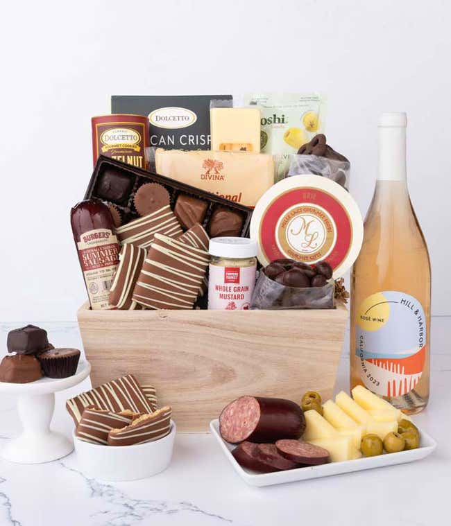 Wooden crate filled with gourmet snacks and a bottle of ros&eacute; wine, including summer sausage, creamy cheeses, chocolate-covered grahams and almonds, assorted chocolates, wafers, crisps, mustard, and olives, styled with plates of cheese and sausage.