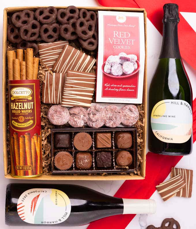 A gift box with chocolate pretzels, chocolate graham crackers, red velvet cookies, hazelnut wafer rolls, an 8 piece box of chocolates and 2 bottles of wine.