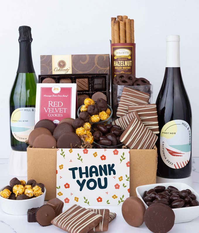 A gift box overflowing with treats including an assortment of chocolate-covered items like oreoes, almonds graham crackers and pretzels along with red velvet cookies, hazelnut wafers, an 8 piece box of chocolates, 2 bottles of wine and a Thank You card.