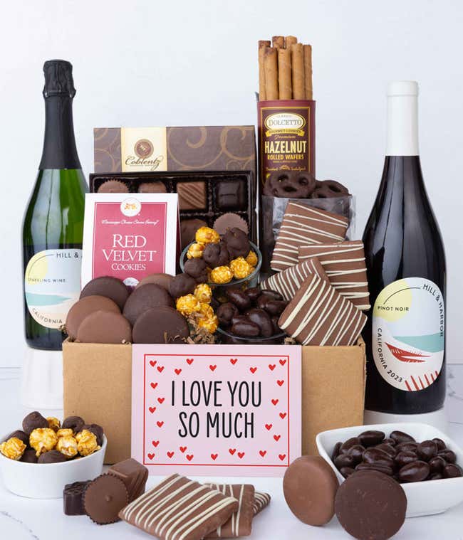 A gift box overflowing with treats including an assortment of chocolate-covered items like oreoes, almonds graham crackers and pretzels along with red velvet cookies, hazelnut wafers, an 8 piece box of chocolates, 2 bottles of wine and an I Love You card.