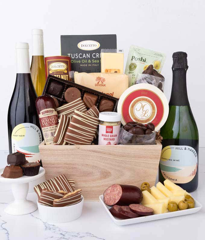 Wooden crate filled with gourmet FOOD and 3 bottles of wine, including Chardonnay, Pinot Noir, and Sparkling Wine, alongside artisan summer sausage, creamy cheeses, assorted chocolates, striped chocolate grahams, cookies, crisps, mustard, and olives.