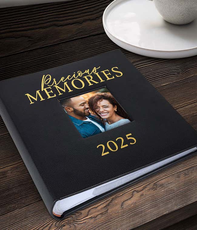Black photo album titled 'Precious Memories 2025' with gold lettering and a square photo inset of a smiling couple, resting on a dark wooden surface near a white tray with a cup.