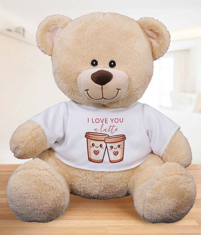 Light tan plush teddy bear sitting on a wooden surface, wearing a white T-shirt that reads &lsquo;I love you a latte&rsquo; with an illustration of two smiling coffee cups.