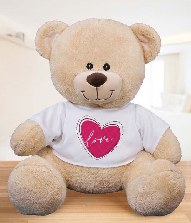 Light tan plush teddy bear sitting on a wooden surface, wearing a white T-shirt with a pink heart design and the word love printed in the center.
